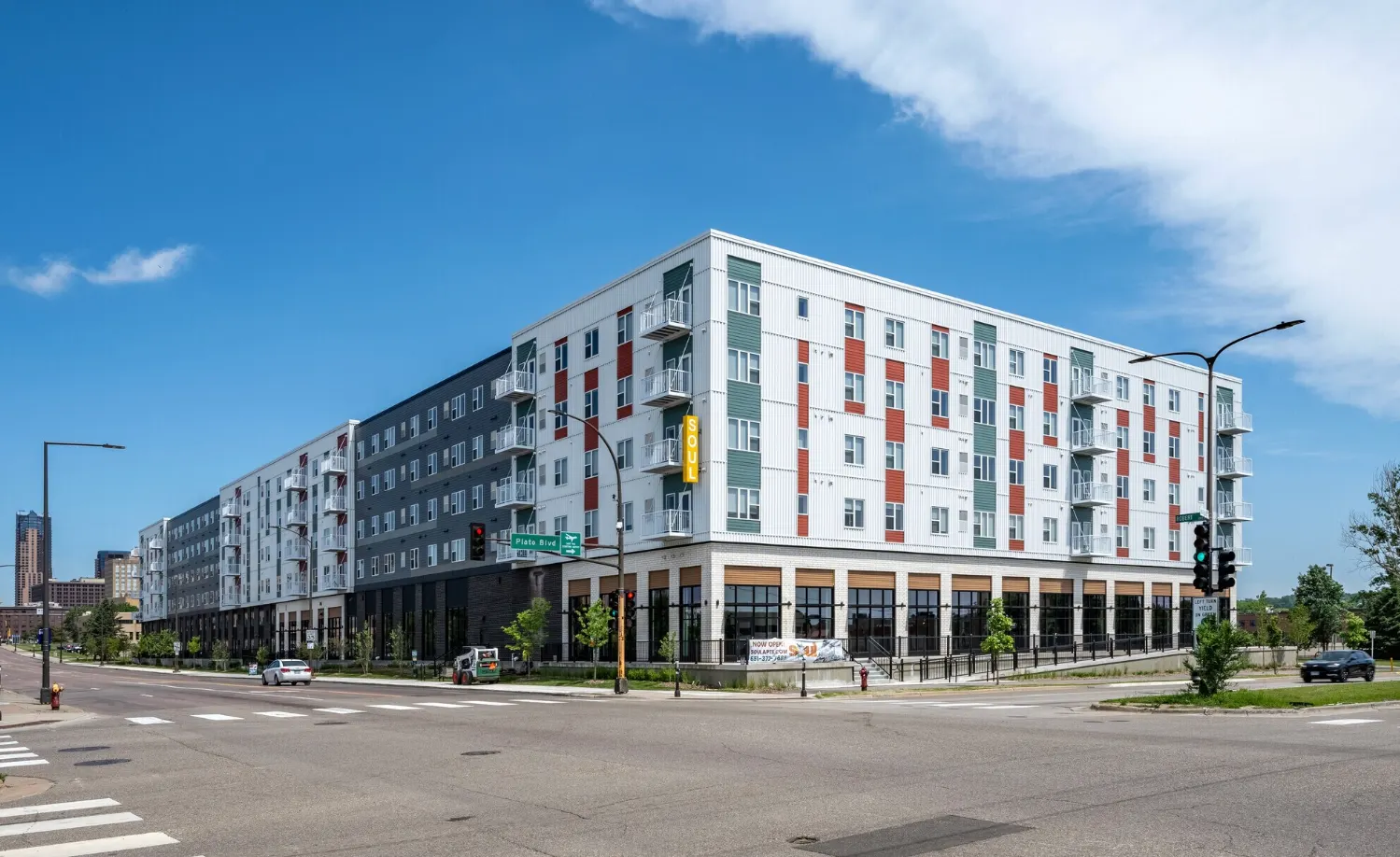 Six-story mixed-use affordable housing development at a signalized urban intersection, featuring ground-floor retail with storefront glazing, residential units with balconies above, and mixed-material cladding including corrugated metal panels with red and green accent sections, representative of LIHTC-financed multifamily construction in a transit-accessible urban infill location.