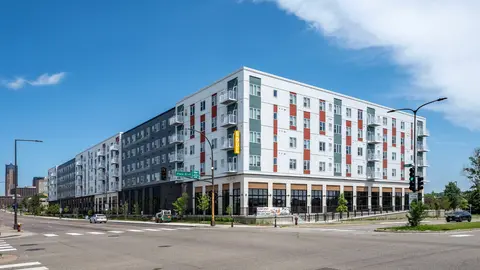 Six-story mixed-use affordable housing development at a signalized urban intersection, featuring ground-floor retail with storefront glazing, residential units with balconies above, and mixed-material cladding including corrugated metal panels with red and green accent sections, representative of LIHTC-financed multifamily construction in a transit-accessible urban infill location.
