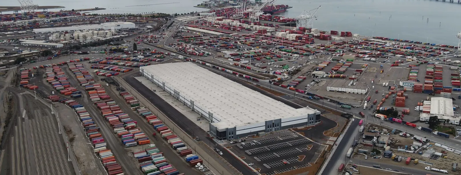 Aerial view of a U.S. port with stacked shipping containers, cranes, docked cargo ships, and a large logistics warehouse near the waterfront.