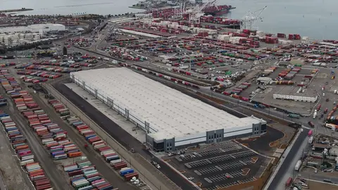 Aerial view of a U.S. port with stacked shipping containers, cranes, docked cargo ships, and a large logistics warehouse near the waterfront.