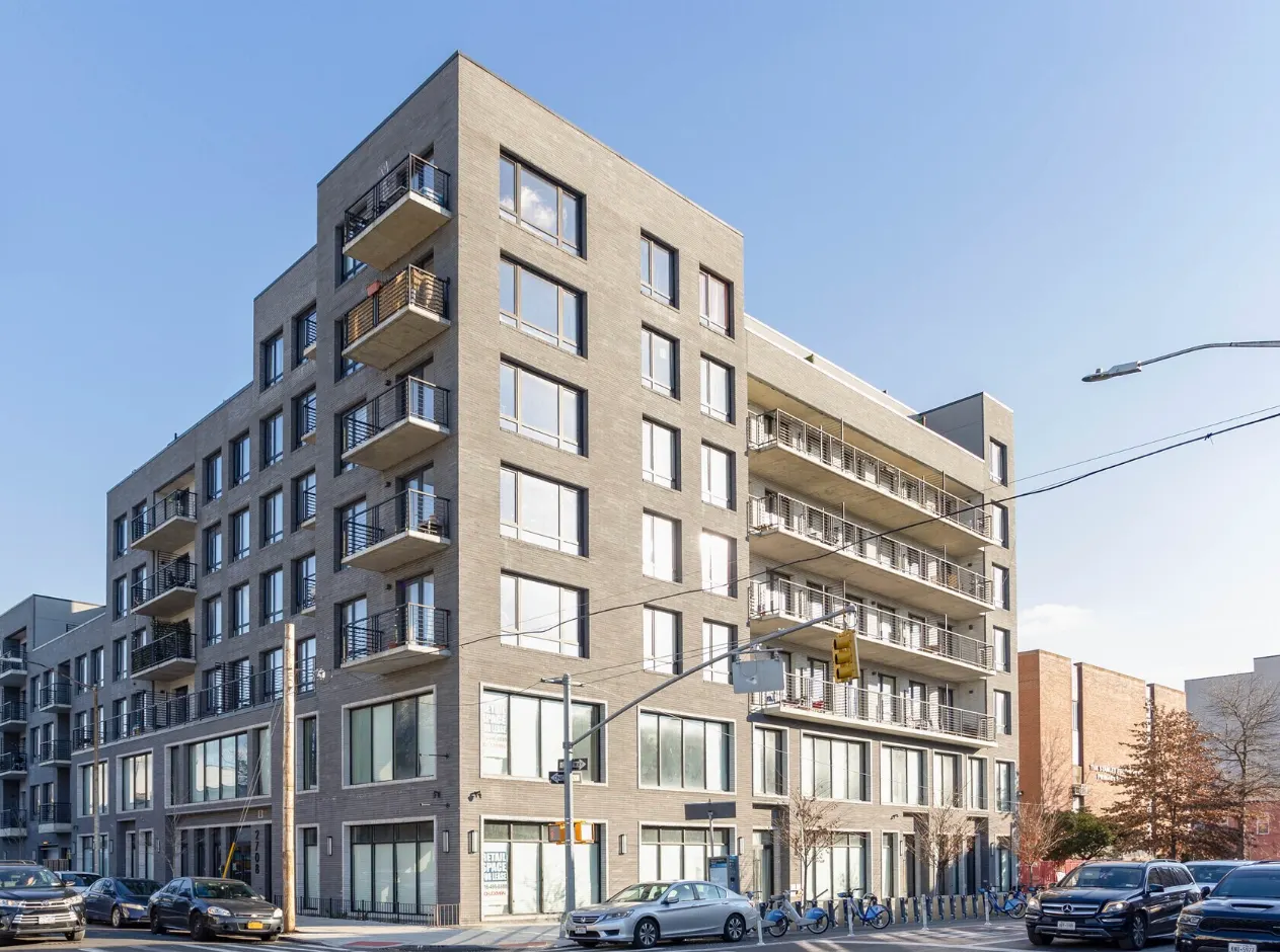 Seven-story mixed-use multifamily building with ground-floor retail, light gray brick facade, cantilevered balconies, and large aluminum-framed windows, located at an urban corner lot, representative of mid-rise infill multifamily development typical of high-density urban-fringe markets.