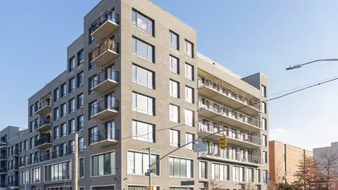 Seven-story mixed-use multifamily building with ground-floor retail, light gray brick facade, cantilevered balconies, and large aluminum-framed windows, located at an urban corner lot, representative of mid-rise infill multifamily development typical of high-density urban-fringe markets.