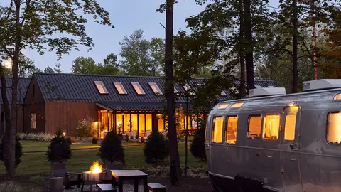 Vintage silver travel trailer and a barn-style lodge at a glamping resort at dusk
