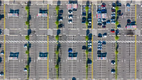 Aerial view of a large parking lot with scattered cars, tree-lined medians, covered cart stations, and marked crosswalks. Most cars are clustered in the upper right area.