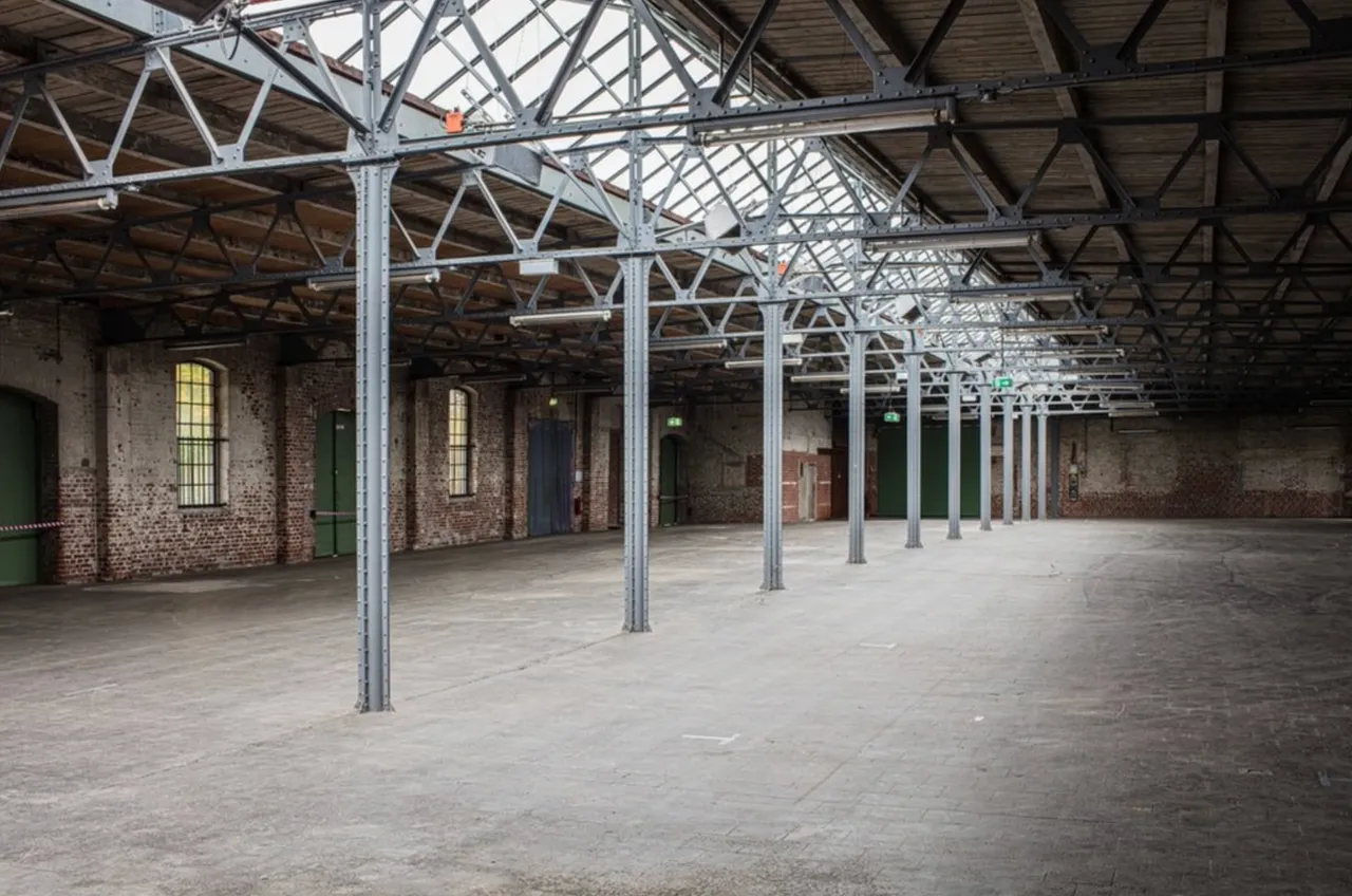 Empty historic industrial hall with steel roof trusses, brick walls and arched windows, before conversion